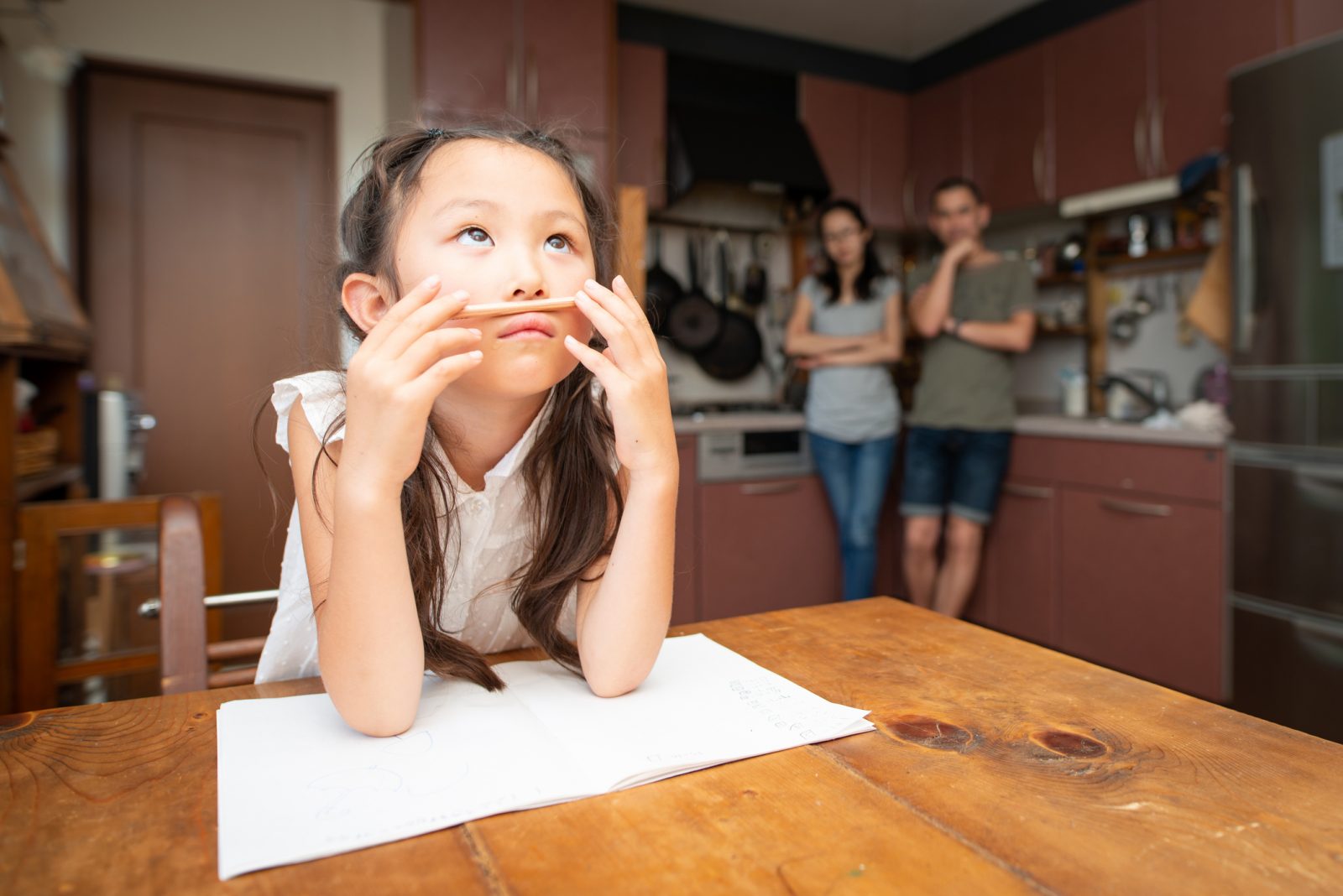A child balances a pencil between her nose and lip while struggling with homework at the kitchen table, while parents watch from the background.