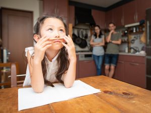 A child balances a pencil between her nose and lip while struggling with homework at the kitchen table, while parents watch from the background.