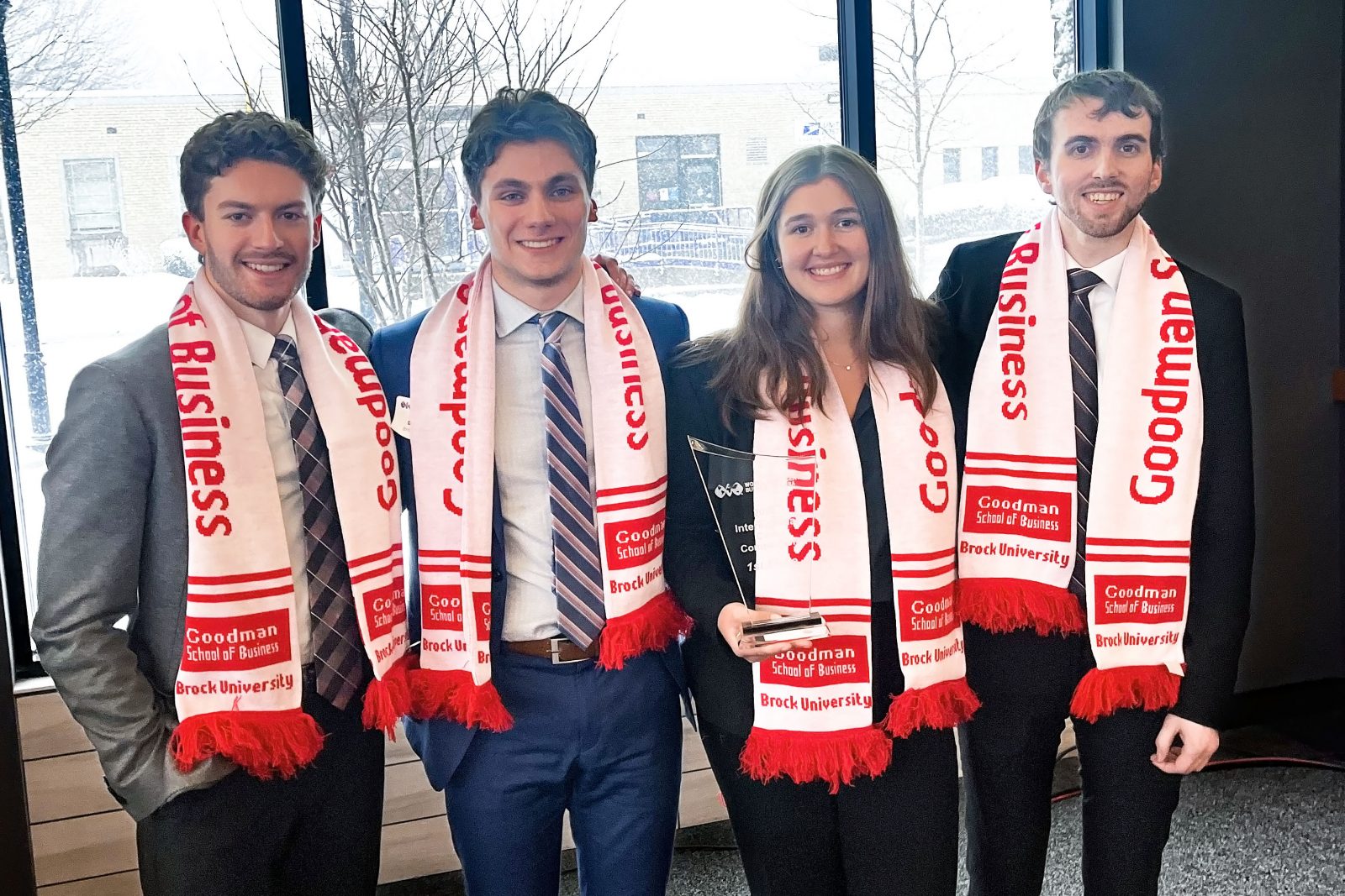 : Four students in suits wear red and white scarves that say Goodman School of Business while holding a clear glass trophy.