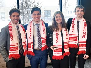 : Four students in suits wear red and white scarves that say Goodman School of Business while holding a clear glass trophy.