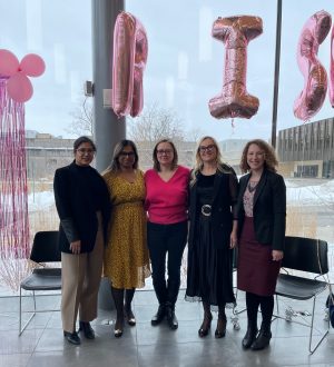 Five women stand in atrium space with pink balloons behind them.