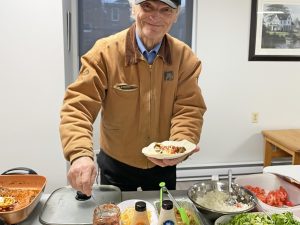 A elderly man serves tacos. An array of taco ingredients can be seen on the table in front of him.