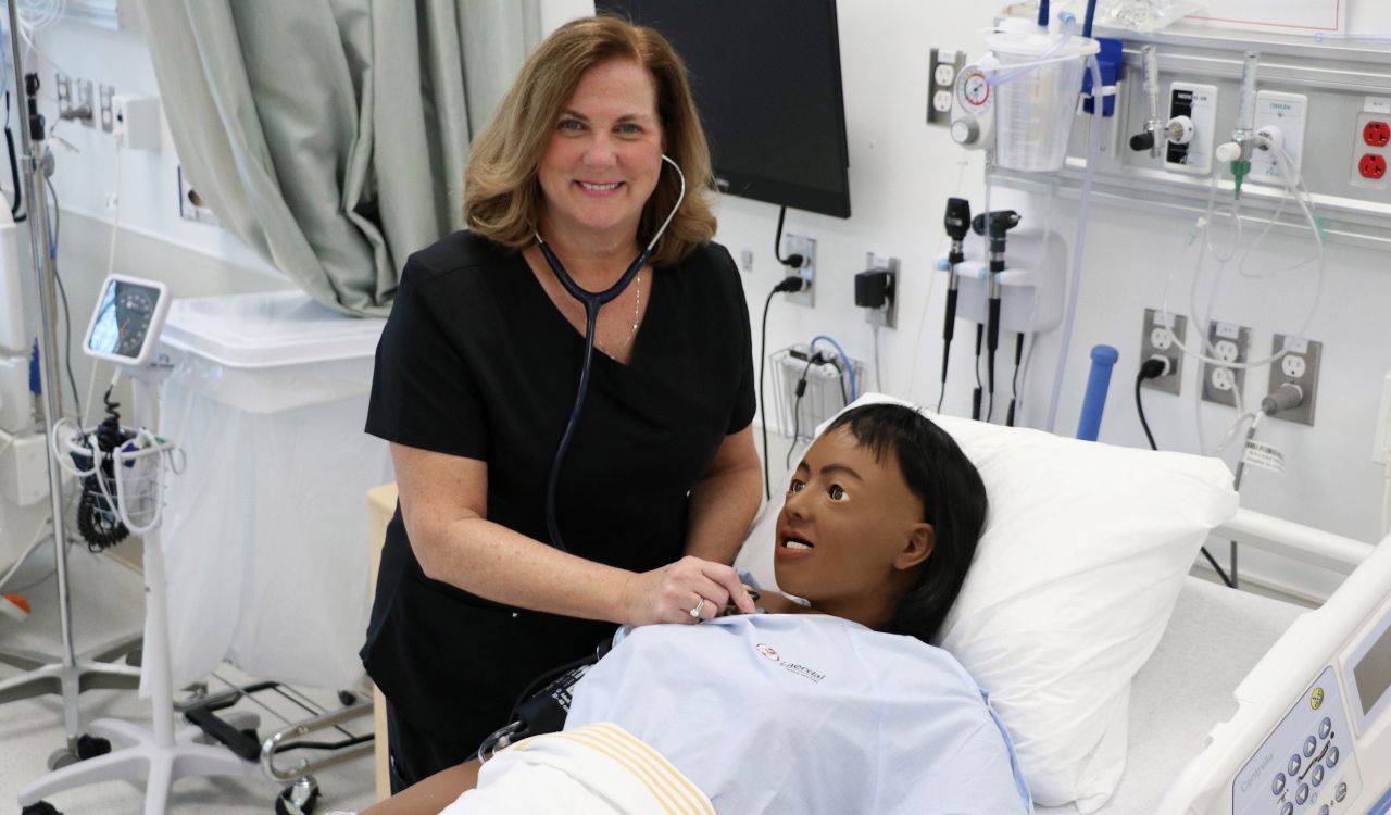 A woman wearing a dark medical uniform leans over a high-fidelity patient simulator to listen to its heartbeat.