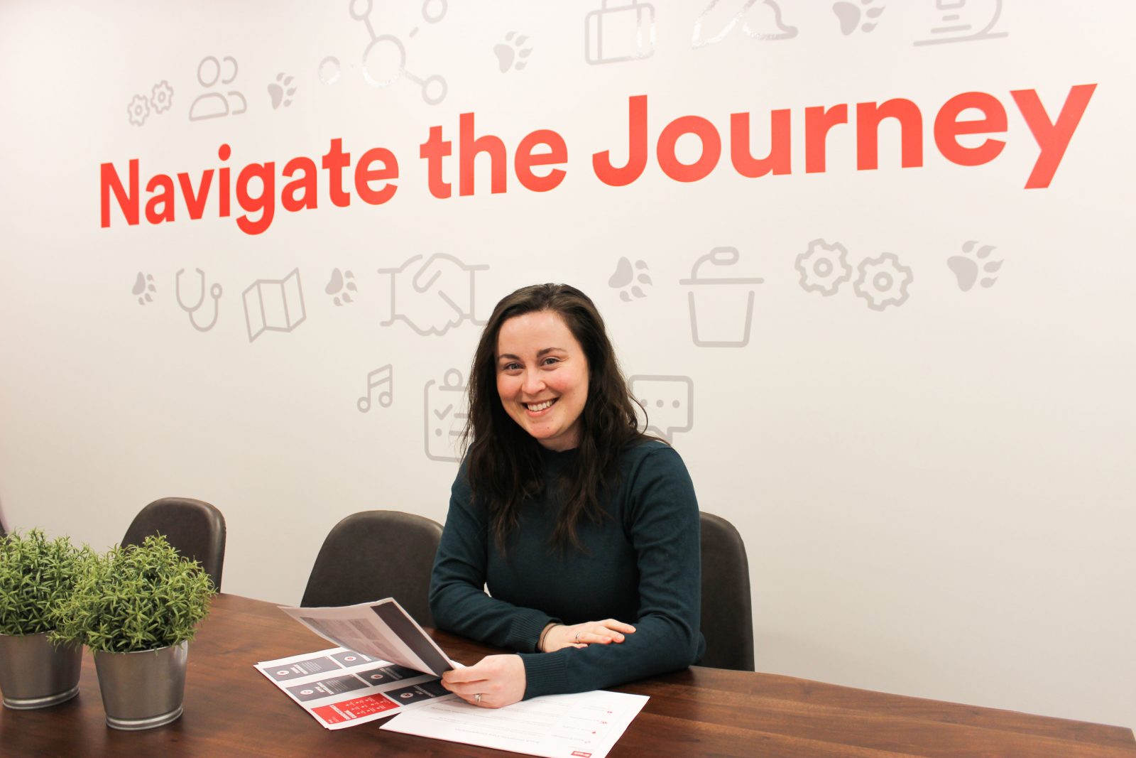 A woman sits at a desk smiling with papers in her hand. A sign on the wall behind her reads 'Navigate the Journey.'