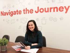 A woman sits at a desk smiling with papers in her hand. A sign on the wall behind her reads 'Navigate the Journey.'