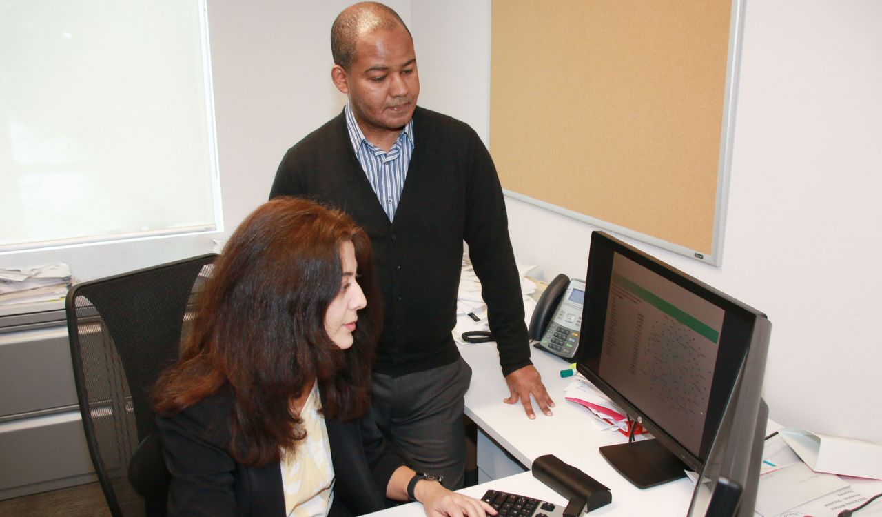 Two people look at computer screens, one seated at a desk and the other standing behind.
