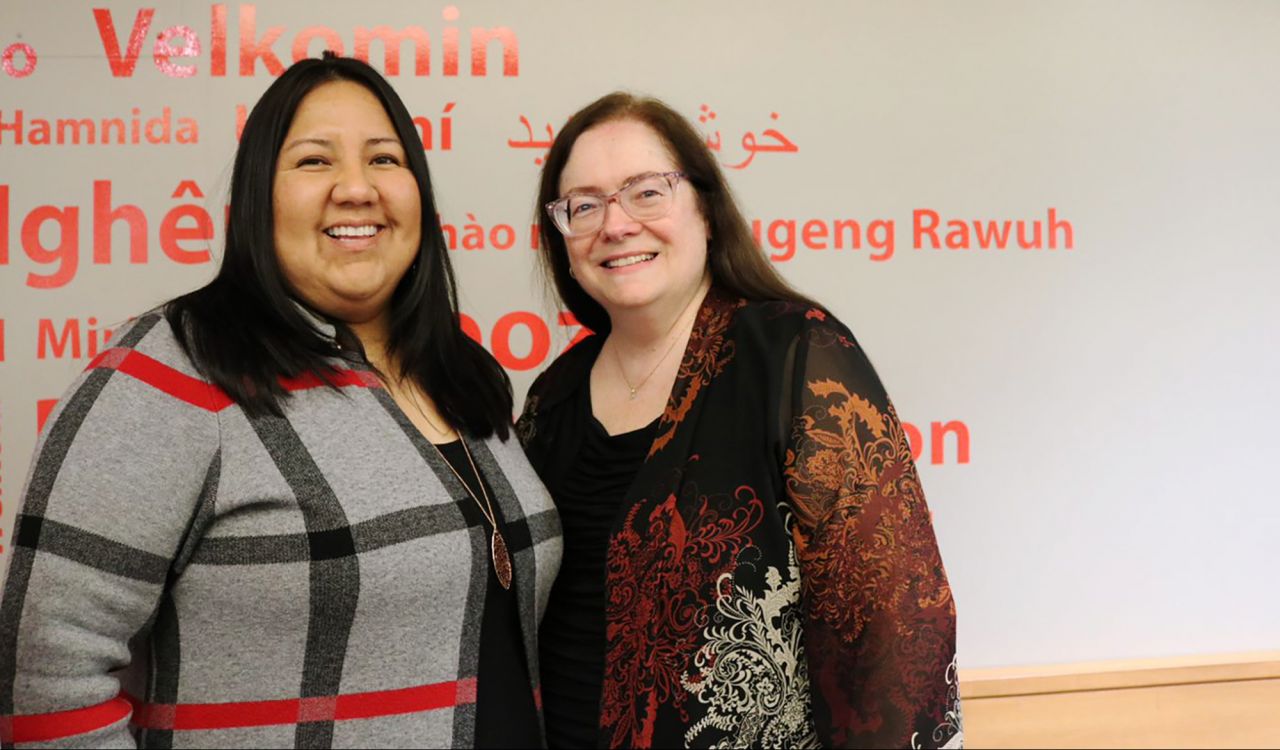 Michelle Bomberry (left) and Sheila O’Keefe-McCarthy (right) with an arm around one another, against a wall with red lettering.
