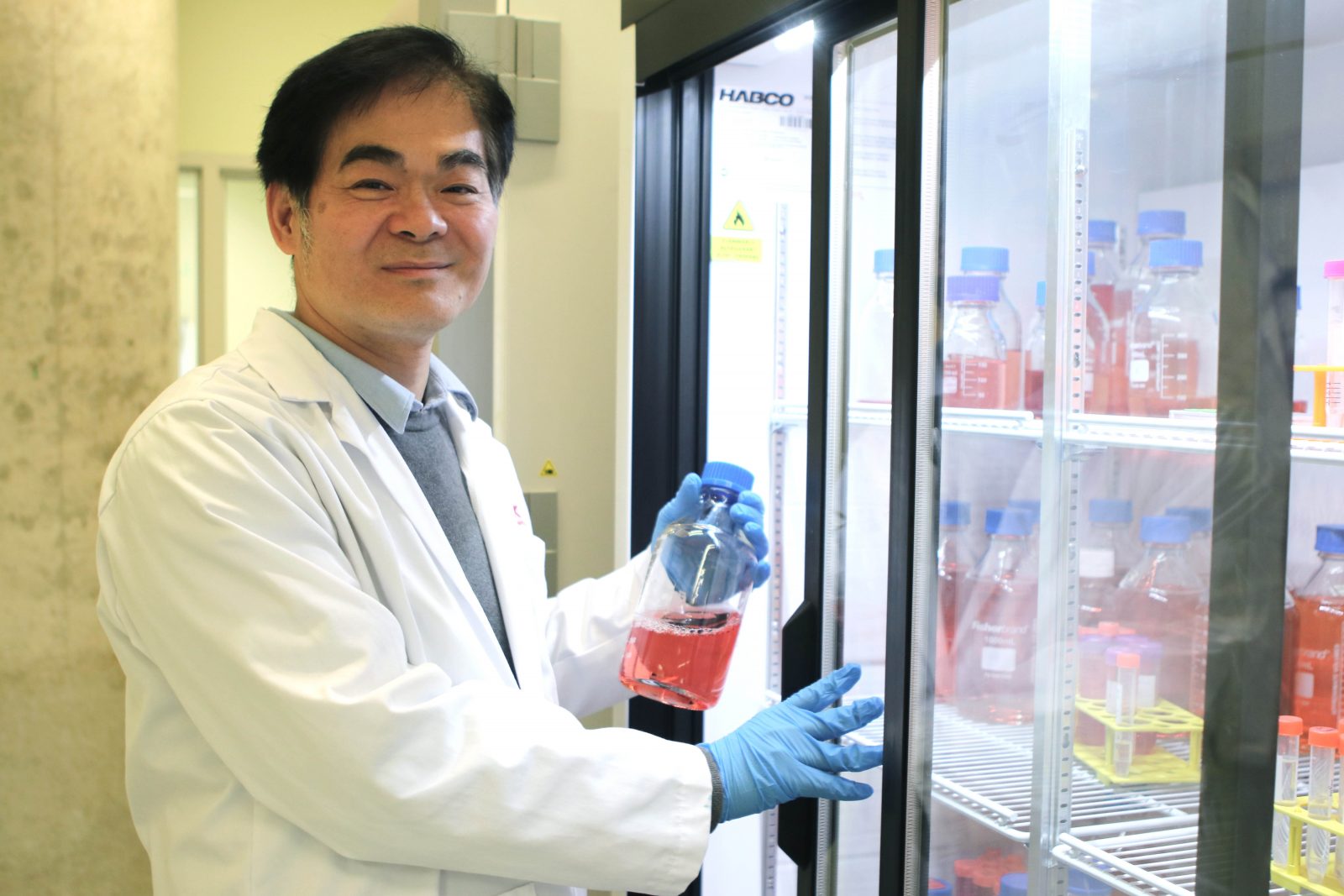 Newman Sze smiles into the camera as he stands next to a refrigerator in his lab.