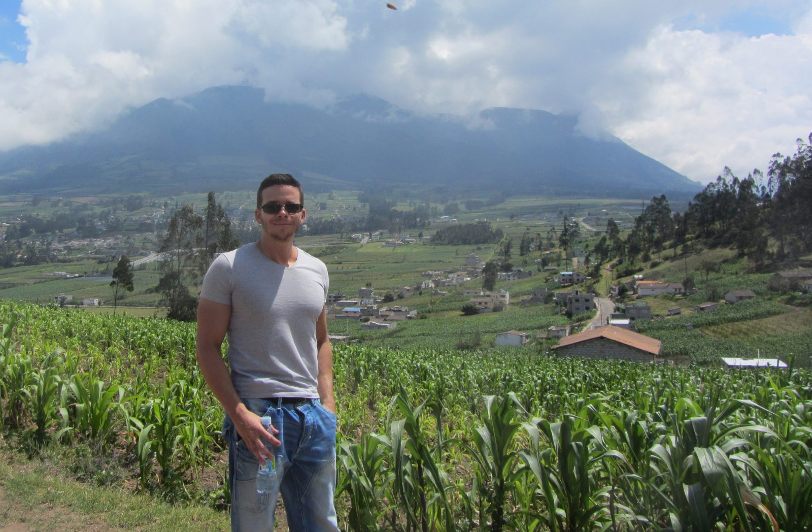 Pascal Lupien stands on a road overlooking a crop field and valley community, with a mountain touching clouds in the background
