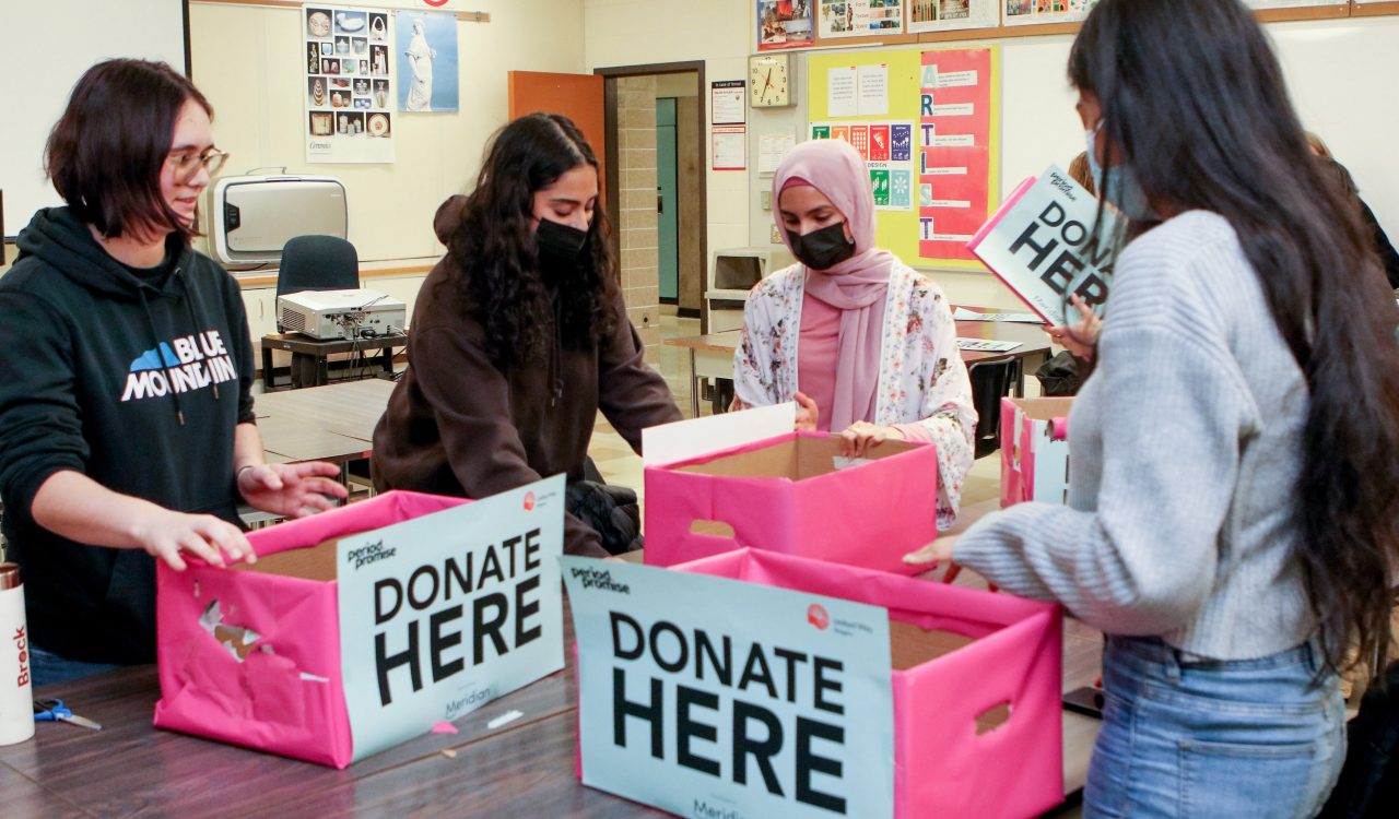 Four students work together to wrap cardboard boxes in pink wrapping paper. Signs on the boxes read ‘donate here.”