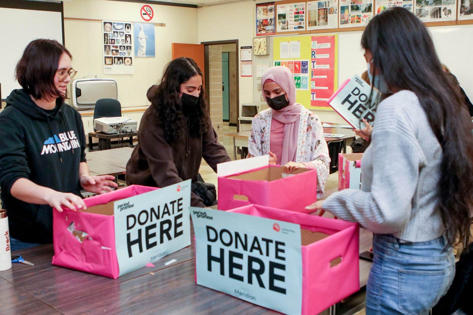 Four students work together to wrap cardboard boxes in pink wrapping paper. Signs on the boxes read ‘donate here.”