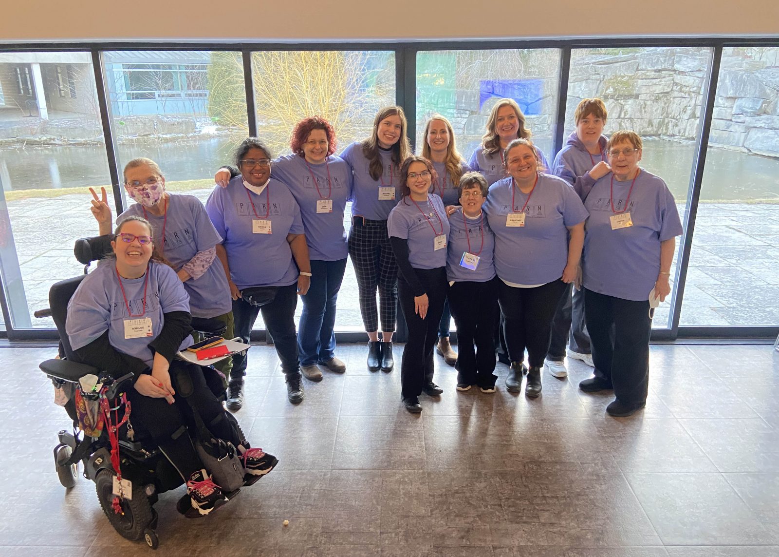 Twelve members of the PARN event planning committee pose in matching purple event t-shirts in front of a wall of windows looking out onto a pond