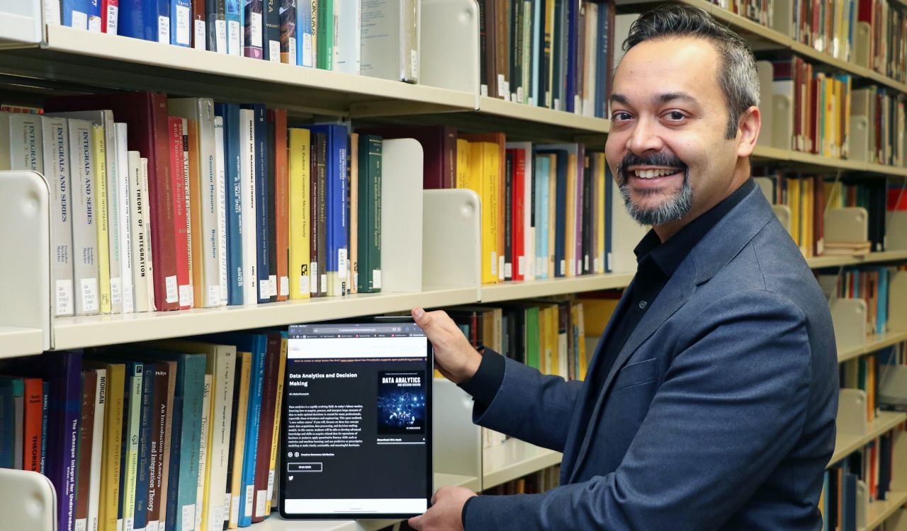Rajiv Jhangiani holds an iPad displaying an open digital textbook as if he is removing it from a library shelf filled with textbooks.