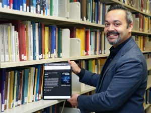 Rajiv Jhangiani holds an iPad displaying an open digital textbook as if he is removing it from a library shelf filled with textbooks.