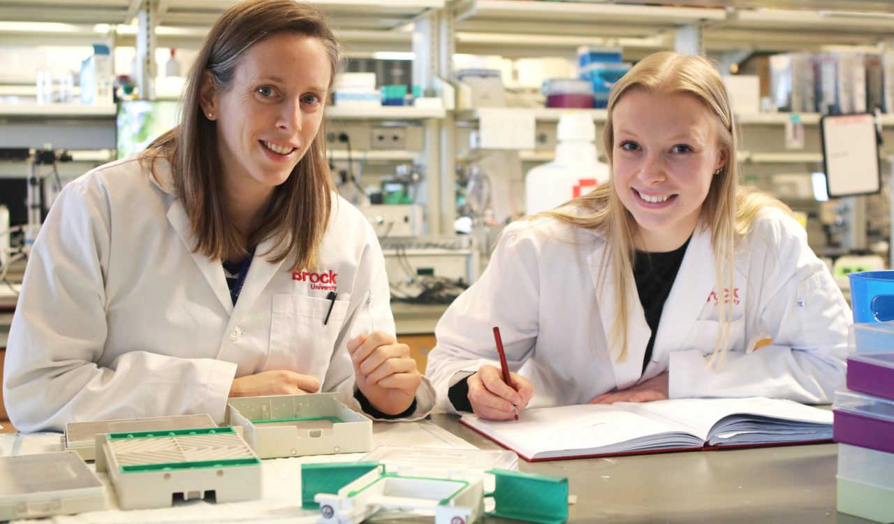 Associate Professor of Health Sciences Rebecca MacPherson and PhD student Emily Copeland sit at a counter in a lab while wearing white lab coats.