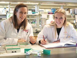 Associate Professor of Health Sciences Rebecca MacPherson and PhD student Emily Copeland sit at a counter in a lab while wearing white lab coats.