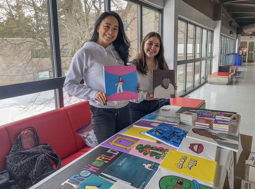 Two female students hold up colourful paintings in front of a table filled with more paintings.