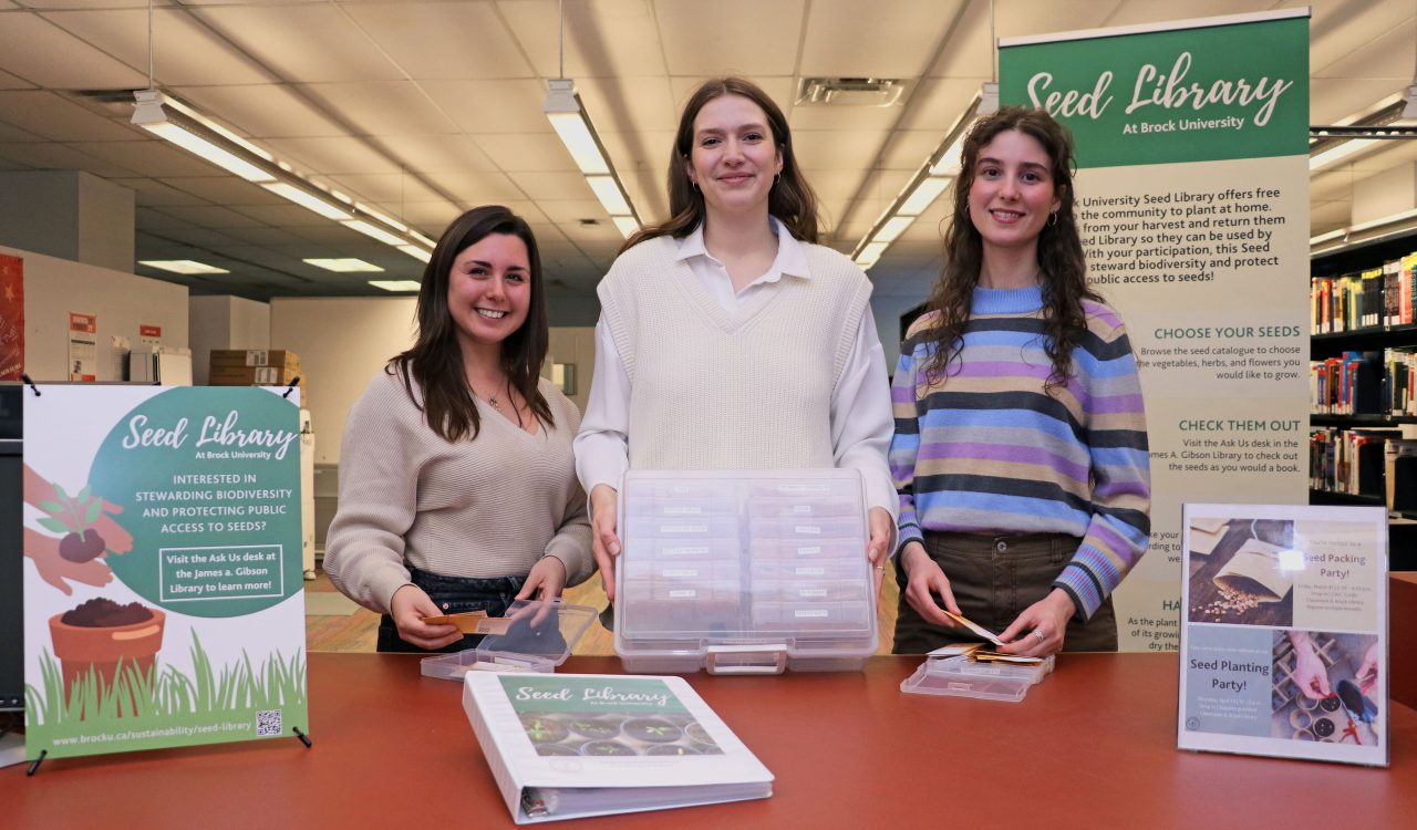 Three women display a catalogue of seeds available in Brock University’s Seed Library.