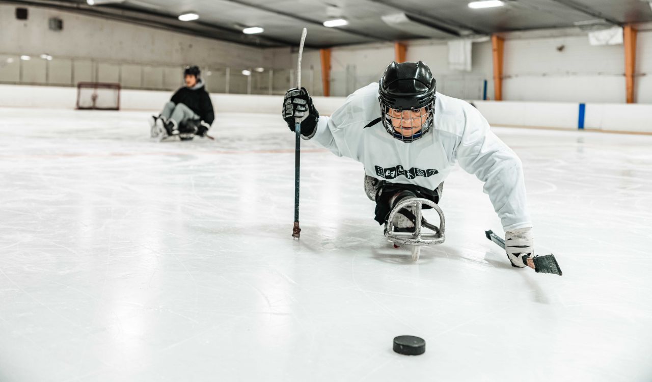 A woman in a hockey helmet and equipment sits on an accessible sledge in the middle of a hockey rink.