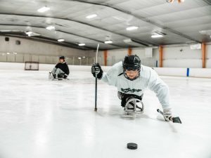 A woman in a hockey helmet and equipment sits on an accessible sledge in the middle of a hockey rink.