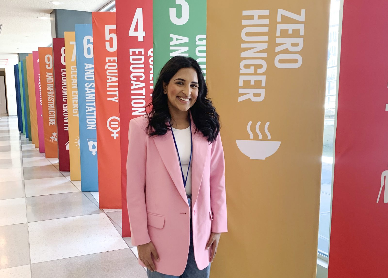 Muskaan Waraich, wearing a pink blazer, stands near a row of banners outlining the UN’s Sustainable Development Goals in a long corridor.