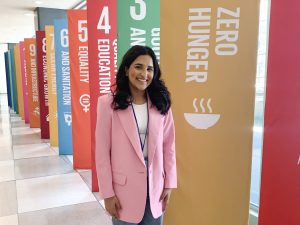 Muskaan Waraich, wearing a pink blazer, stands near a row of banners outlining the UN’s Sustainable Development Goals in a long corridor.