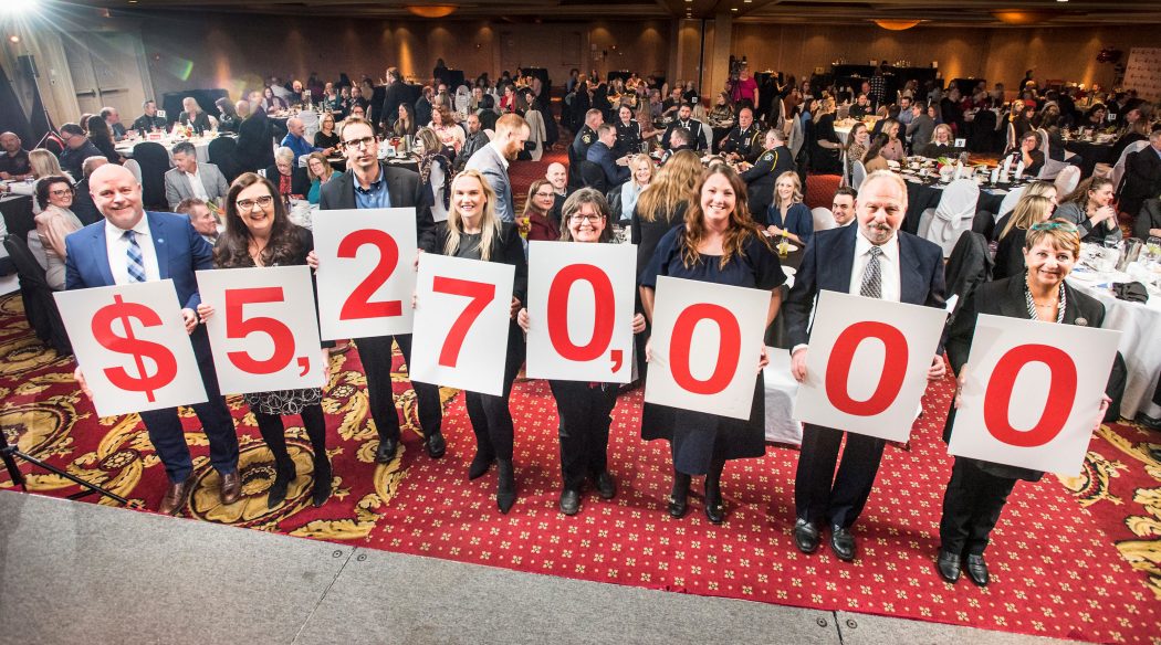 A group of eight people at the front of a large banquet gathering each hold a number on a bristle board, that together reveals $5,270,000. 