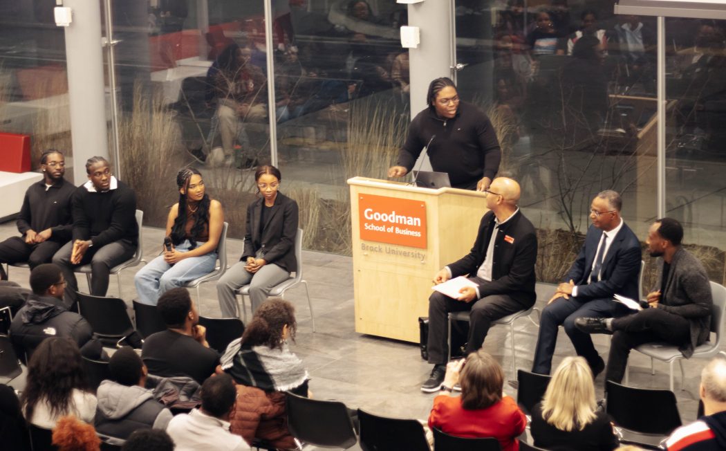 A panel of people sit in front of a crowd having a discussion. A person stands at a podium moderating.