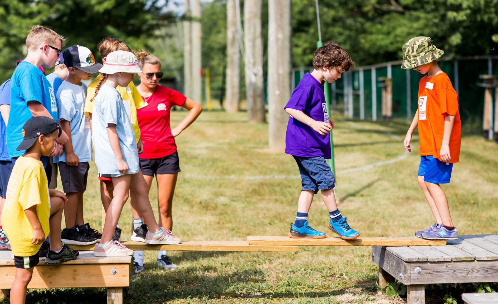 Children walk across a shallow wooden plank during a learning exercise outdoors.