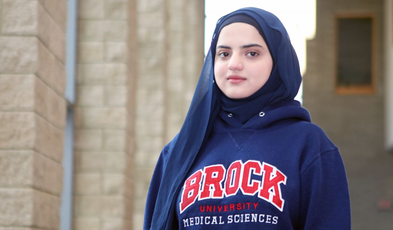 A portrait of a young woman wearing a hijab and a sweater that reads 'Brock University Medical Sciences.'