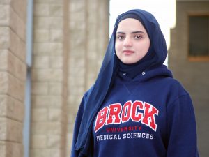 A portrait of a young woman wearing a hijab and a sweater that reads 'Brock University Medical Sciences.'