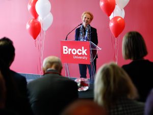 Brock University Chancellor Hilary Pearson stands at a podium flanked by red and white balloons. She is addressing a crowd of people, whose backs can be seen.