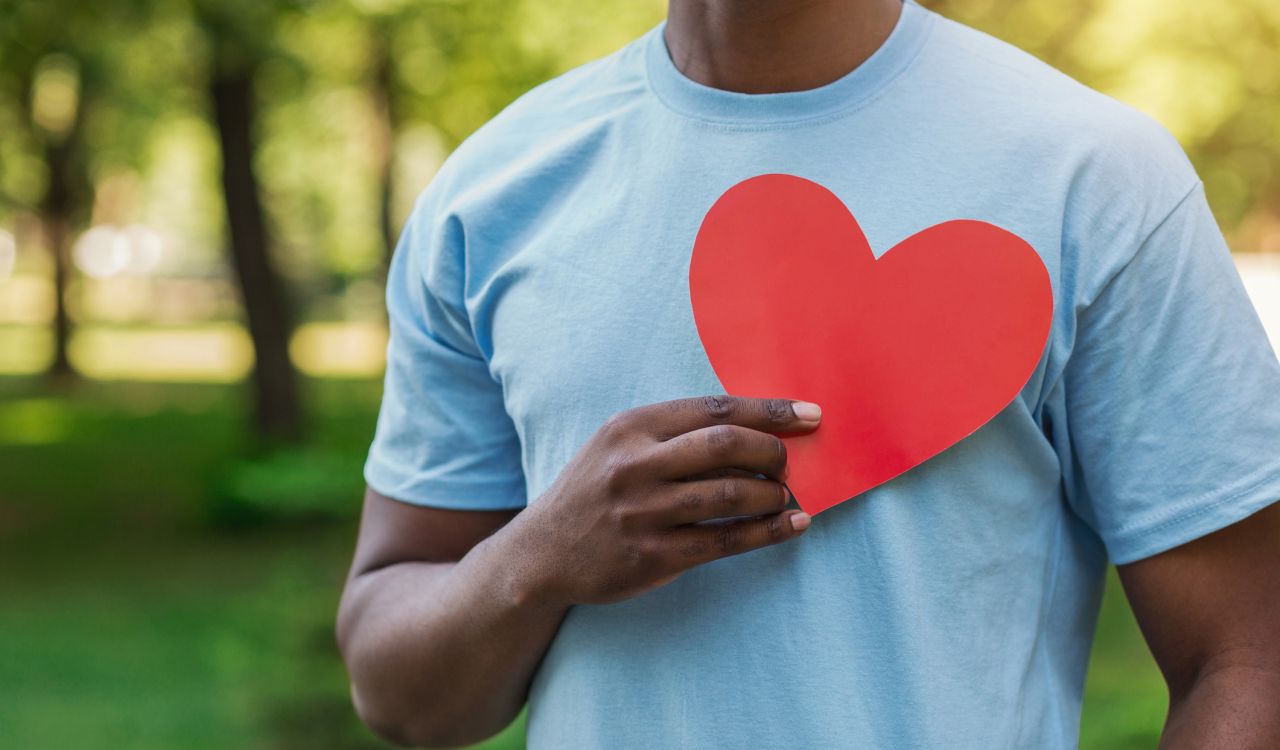 A man in a pale blue t-shirt holds a red paper heart against his chest while standing in a park.