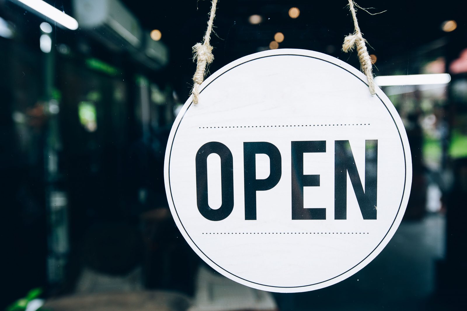 Close-up view of an open sign hanging in a glass door. The sign is round and white with the word "open" printed in black.