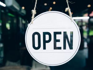 Close-up view of an open sign hanging in a glass door. The sign is round and white with the word "open" printed in black.