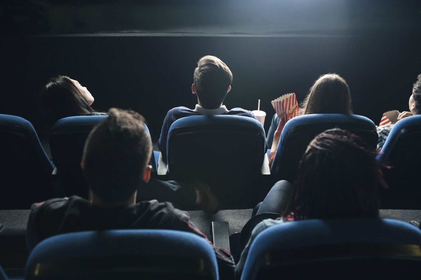 People sitting with popcorn and drinks in blue theatre seats look up at a movie screen, shown from behind.