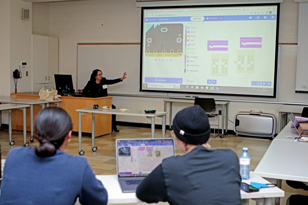 A woman sits at a desk at the front of a classroom and points to a projector screen displaying Micro:bit software. In the foreground of the photo are the backs of two workshop participants, one with their laptop open to the same website displayed on the projector screen. 