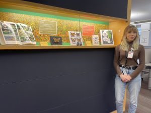A young woman stands in front of an exhibit display case in the Brock Library learning commons. The exhibit features pinned butterflies and moths, as well as books on the subject of pollination.