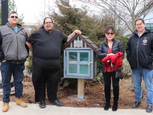 A group of four people stand in line beside a wooden little library. 