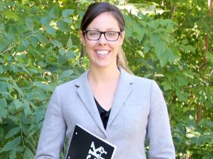 Jessica Blythe stands in front of foliage wearing a grey blazer and glasses, and clutching a black Brock University notebook in her left hand with her right hand in her pants pocket.