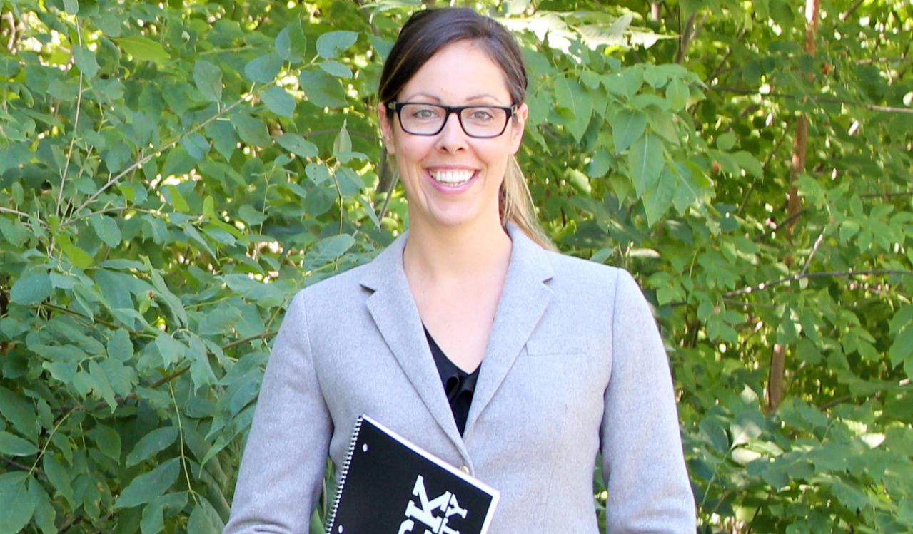 Jessica Blythe stands in front of foliage wearing a grey blazer and glasses, and clutching a black Brock University notebook in her left hand with her right hand in her pants pocket.