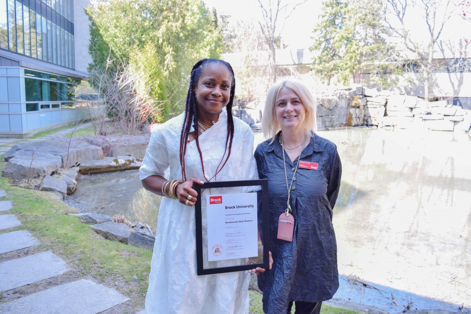 Two women pose as one holds a framed award in front of an outdoor pond.