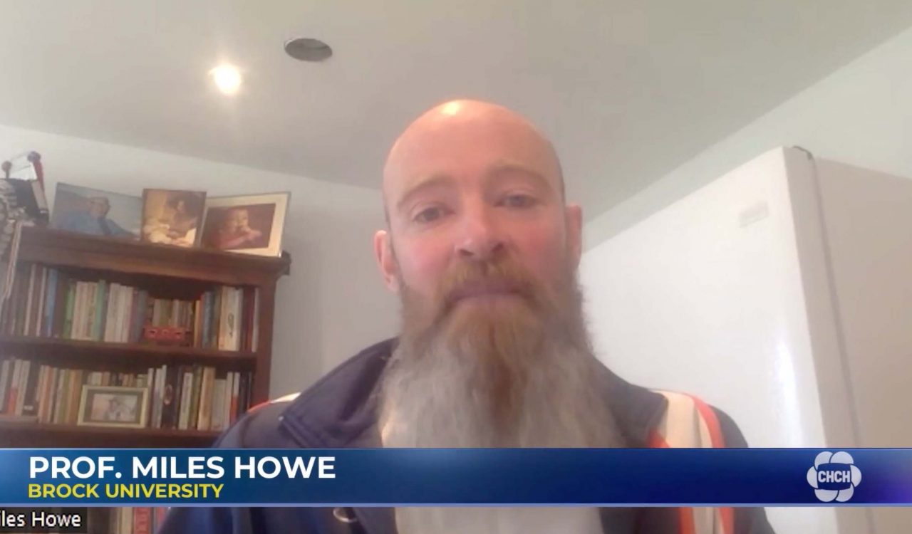 A bearded man speaks to a camera with a bookshelf in the background.
