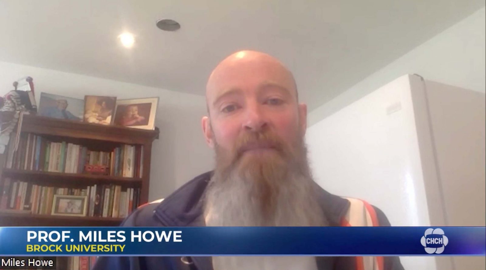A bearded man speaks to a camera with a bookshelf in the background.