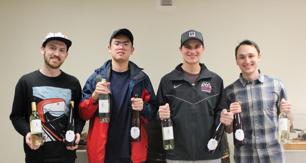 Four students stand holding bottles of wine.