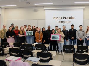 Students gather at the front of a classroom in front of a screen with “Period Promise” projected onto it and pose with pink donation boxes.