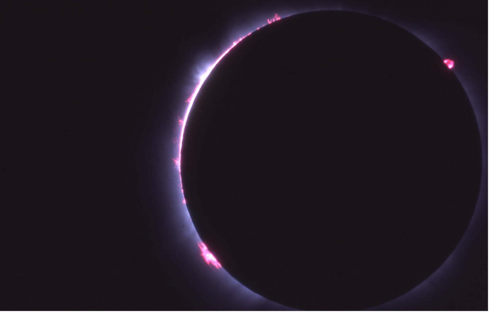 A solar eclipse: the moon is blocking most of the sun. Only a small portion of the sun, glowing pink and purple, can be seen behind the dark shadow of the moon.
