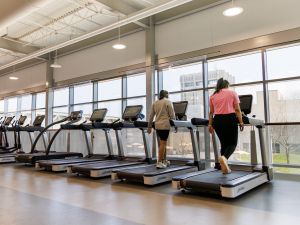 Two women walk on treadmills inside a gym surrounded by natural light.