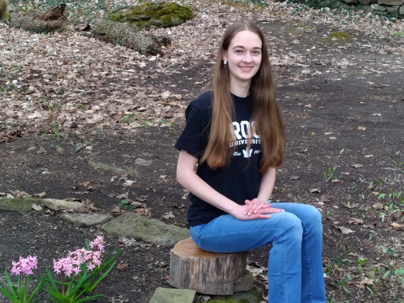 A woman sits on a wooden tree stump in an outdoor setting.