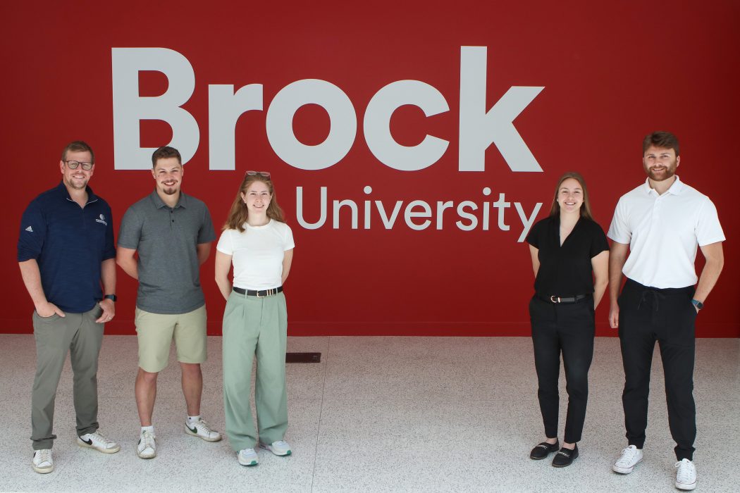 Five people stand next to each other in a line in front of a red wall with the Brock University logo on it. 